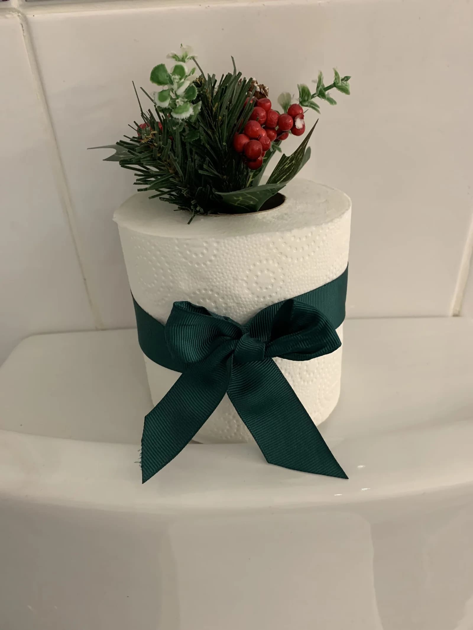 Spare toilet roll tied with a wide green ribbon and a sprig of faux greenery and red berries, standing on a toilet tank