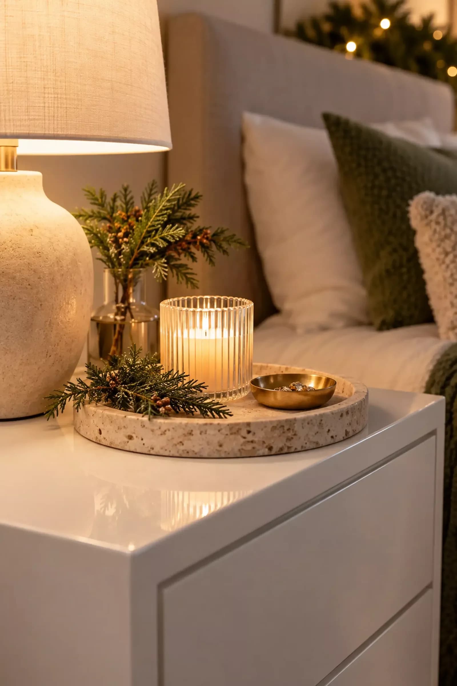 White nightstand with a round stone tray holding a ribbed glass candle, evergreen sprigs in a small vase and a brass dish beside a linen lamp