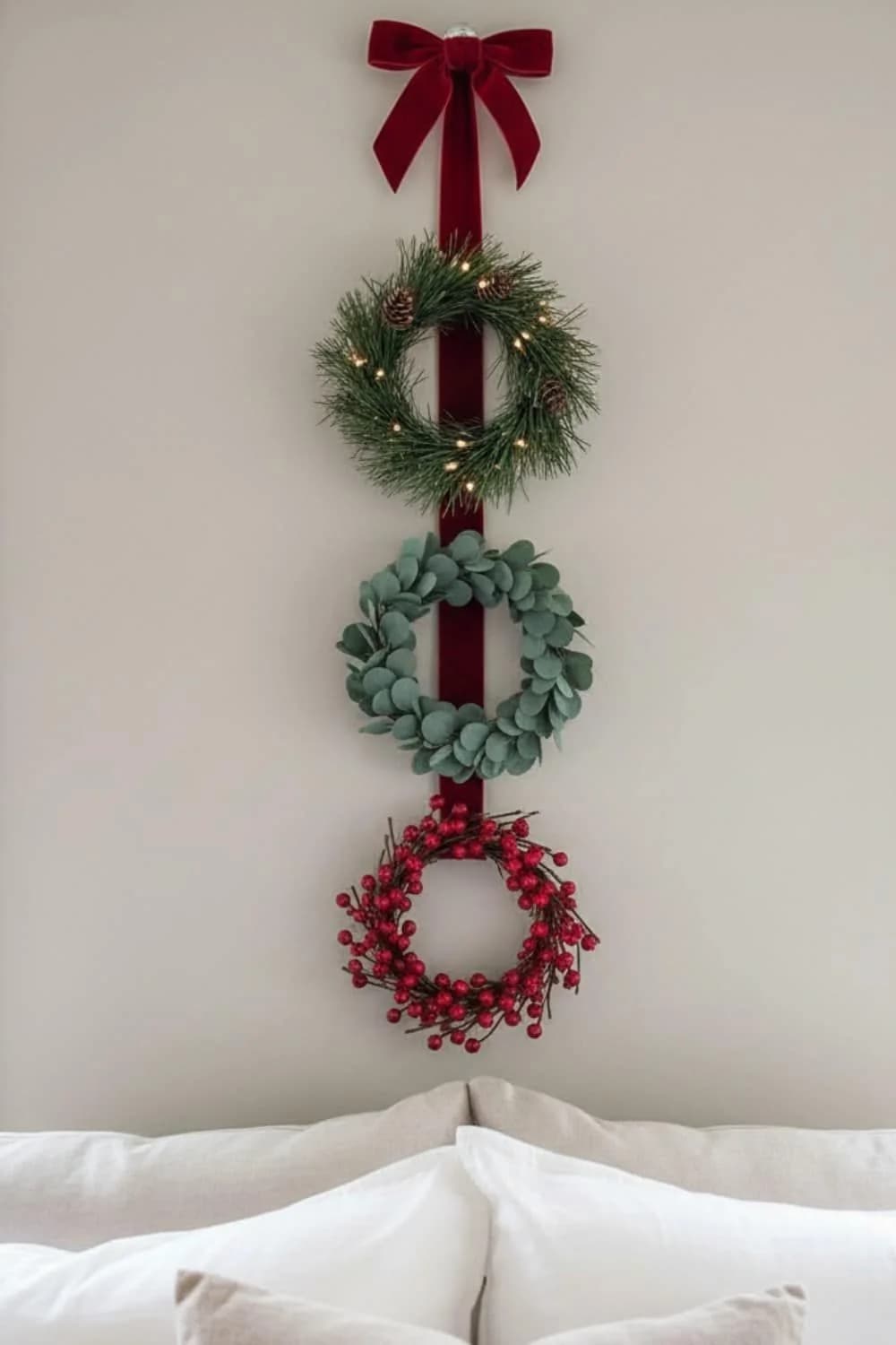 Three small wreaths of pine, eucalyptus and red berries hung in a vertical row on a burgundy velvet ribbon with a bow above a bed
