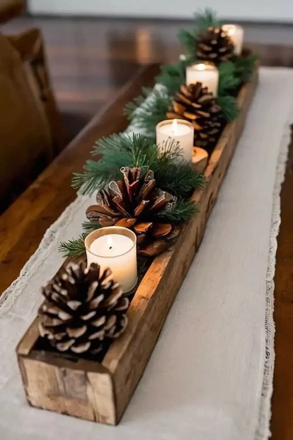 Long rustic wooden box on a linen runner filled with pine, large pine cones and candles in glass holders