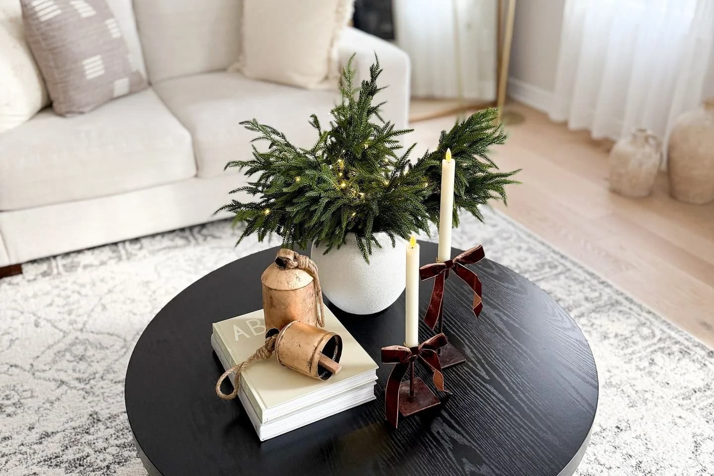 Round black coffee table with a white vase of evergreen branches, two cream taper candles with burgundy bows, and brass bells on a stack of two books, in front of a cream sofa