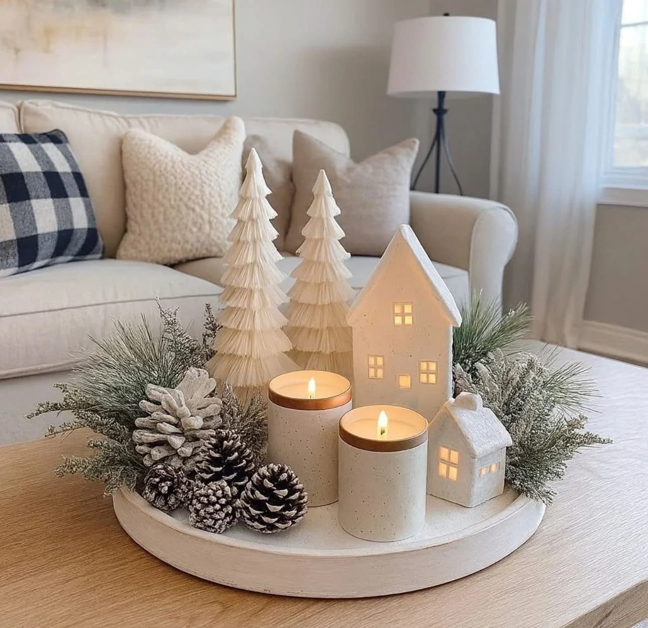 White round tray on a pale wood coffee table with two white paper trees, two white ceramic houses with lit windows, two candles in stone holders, frosted pine cones and greenery