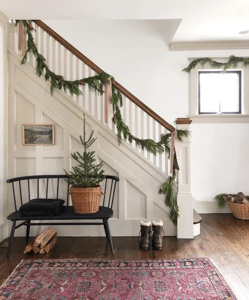 White paneled staircase with an evergreen garland looped along the rail, a black bench holding a small tree in a basket, boots and a patterned rug