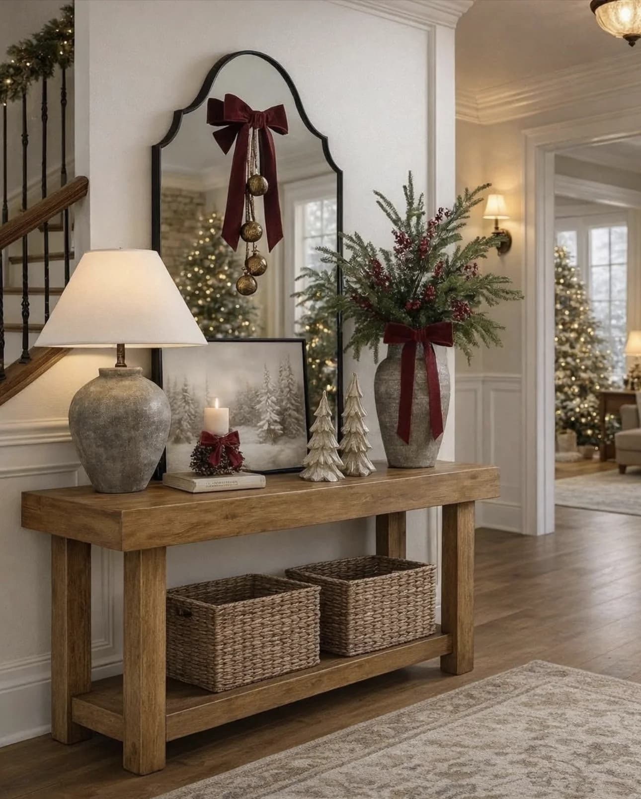 Wooden console with a stone lamp, a leaning winter print, small ceramic trees, a vase of greenery with a burgundy bow and two woven baskets on the lower shelf