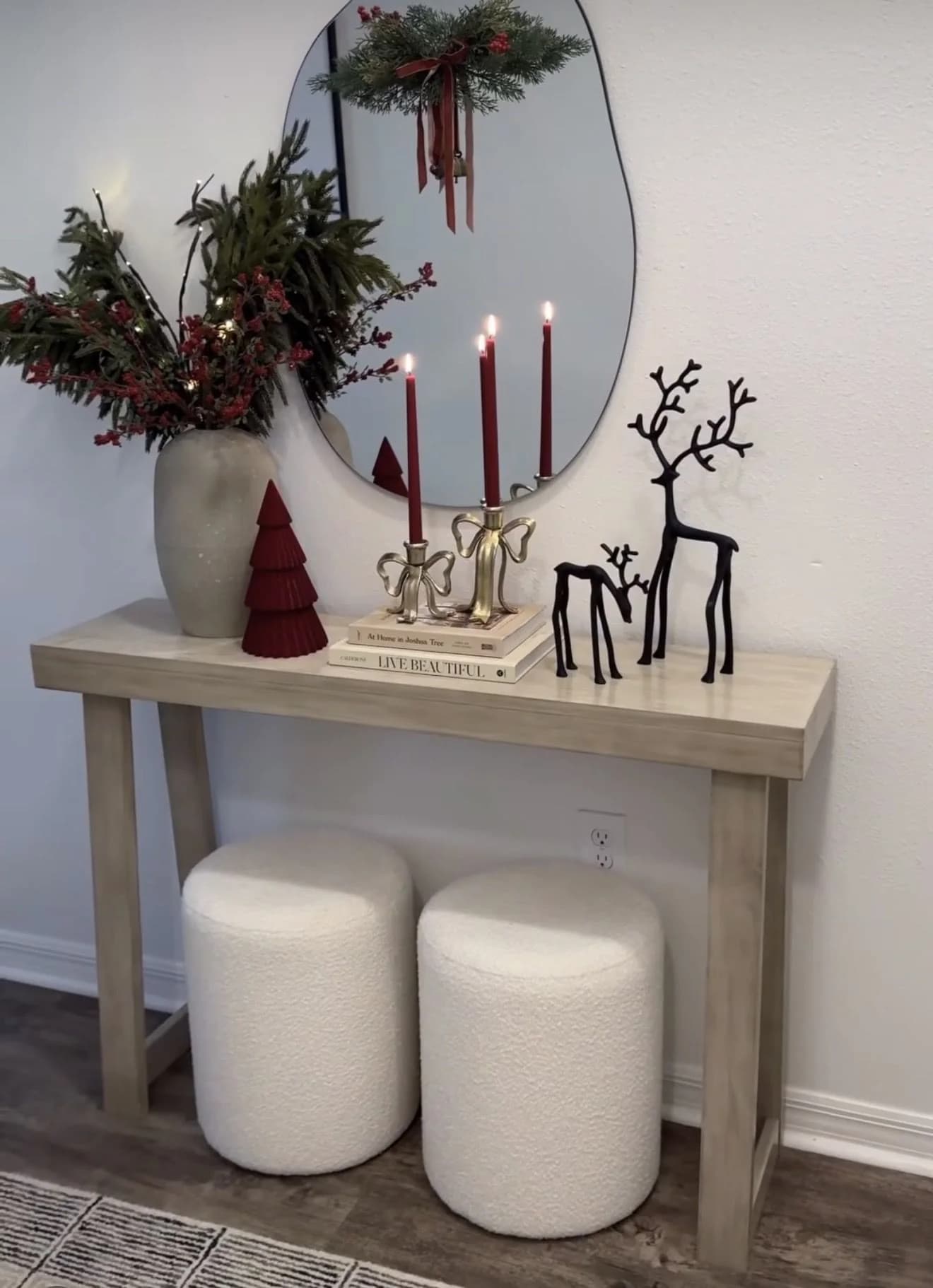 Pale console table with a vase of dark berry branches, burgundy taper candles, black metal reindeer and a swag on an oval mirror above two white stools