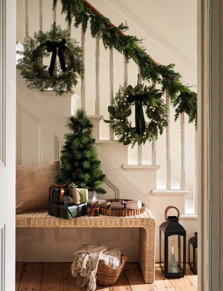 Staircase with two wreaths tied with dark bows on the spindles and a garland on the rail above a woven bench with a small tree, gifts and a lantern