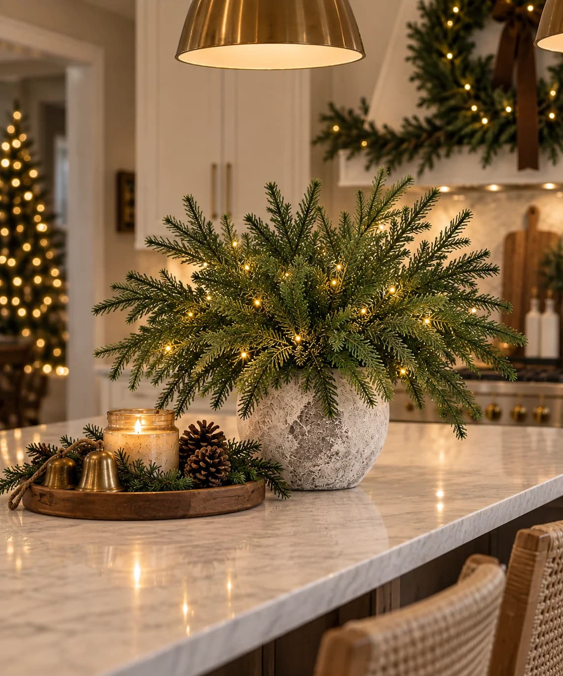 Stone vase of evergreen branches wound with micro lights on a marble island, with a small wooden tray of pine cones, brass bells and a candle beside it