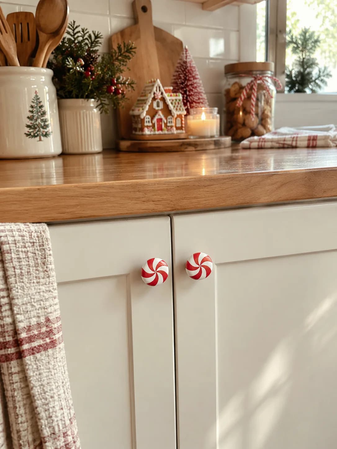 Close view of cream cabinet doors with two red and white peppermint knobs, a wood counter above with a gingerbread house, a candle, a crock of wooden spoons with a tree motif and a red checked towel
