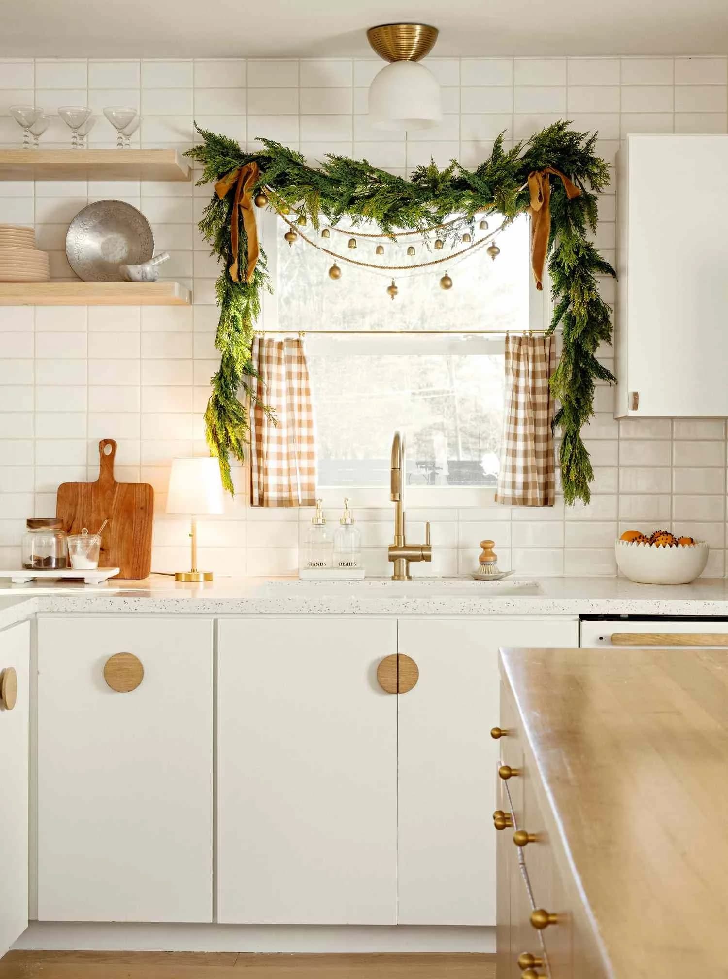 Window over a kitchen sink with a short evergreen swag tied with tan ribbons, a string of small brass bells, tan gingham cafe curtains and a small lamp on the counter