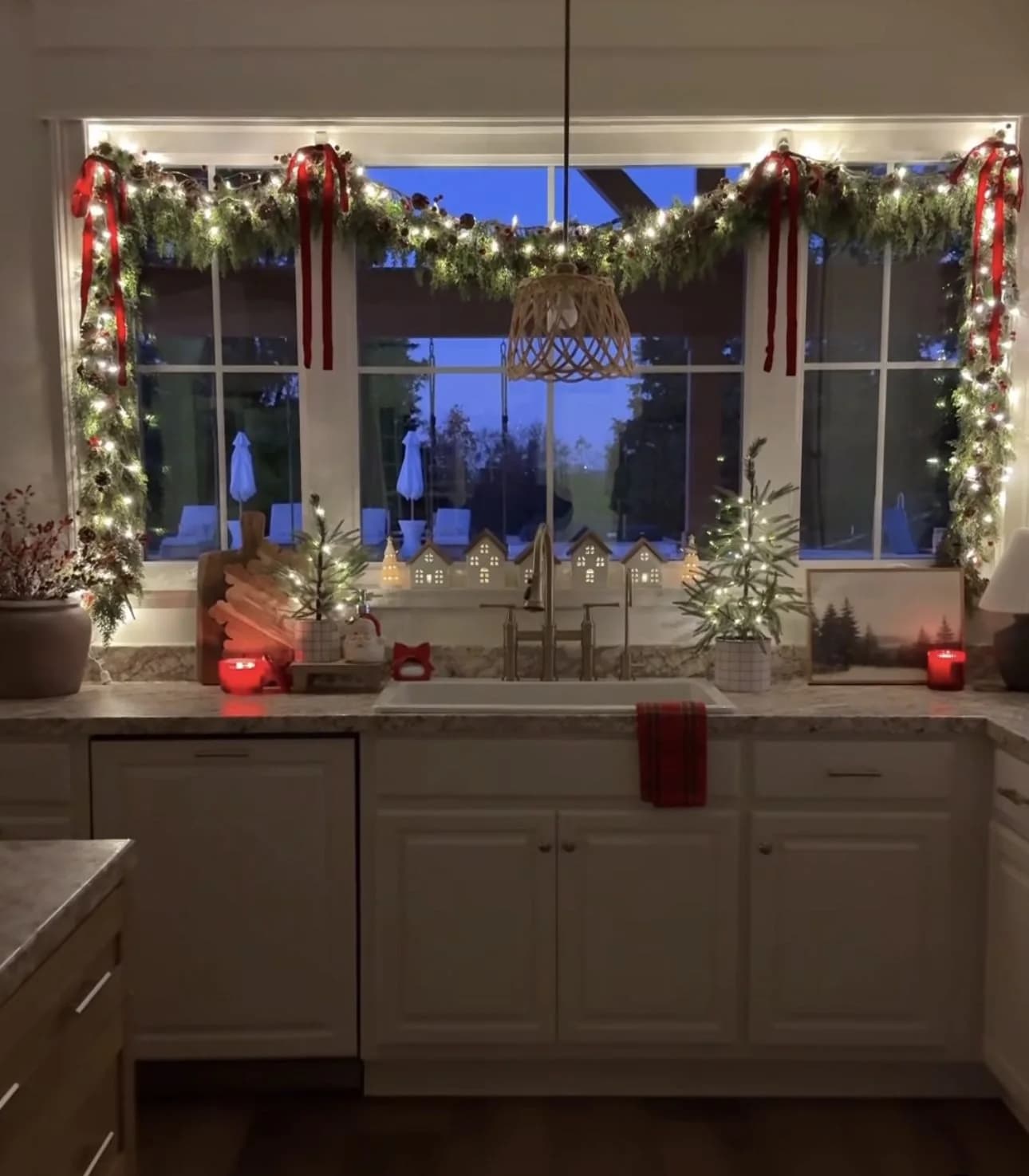 Wide kitchen window at dusk with a lit garland and red ribbons across the top, a row of small lit houses and two small lit trees on the sill, and a red towel on the cabinet below