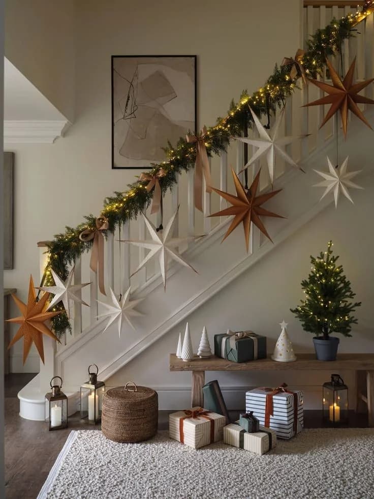 Staircase with a lit garland along the rail and large white and rust paper stars hanging on the open side, a bench with wrapped gifts, lanterns and a small tree below