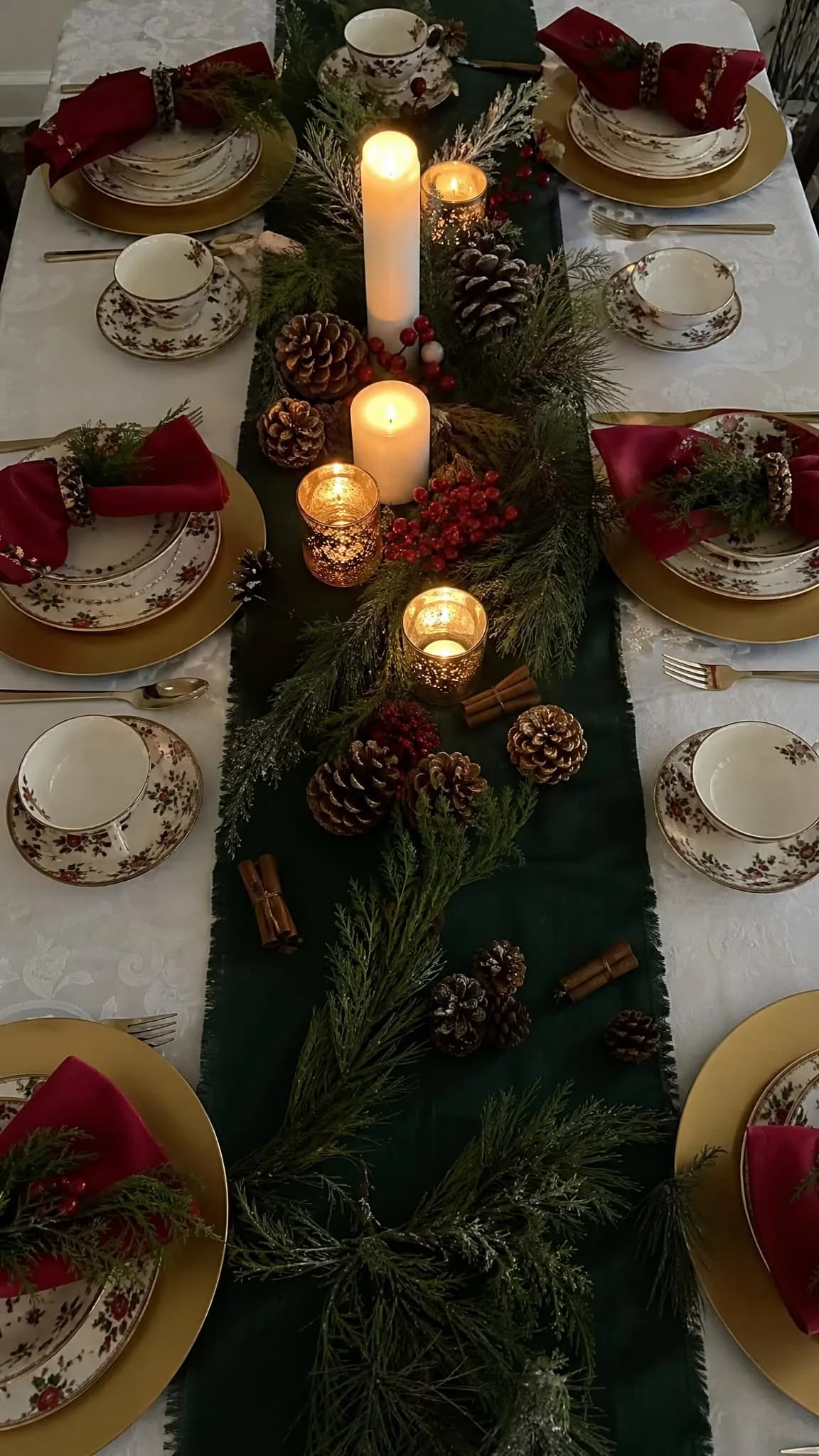 Overhead view of a table with a dark green fabric runner, pine branches, pine cones, cinnamon sticks and candles