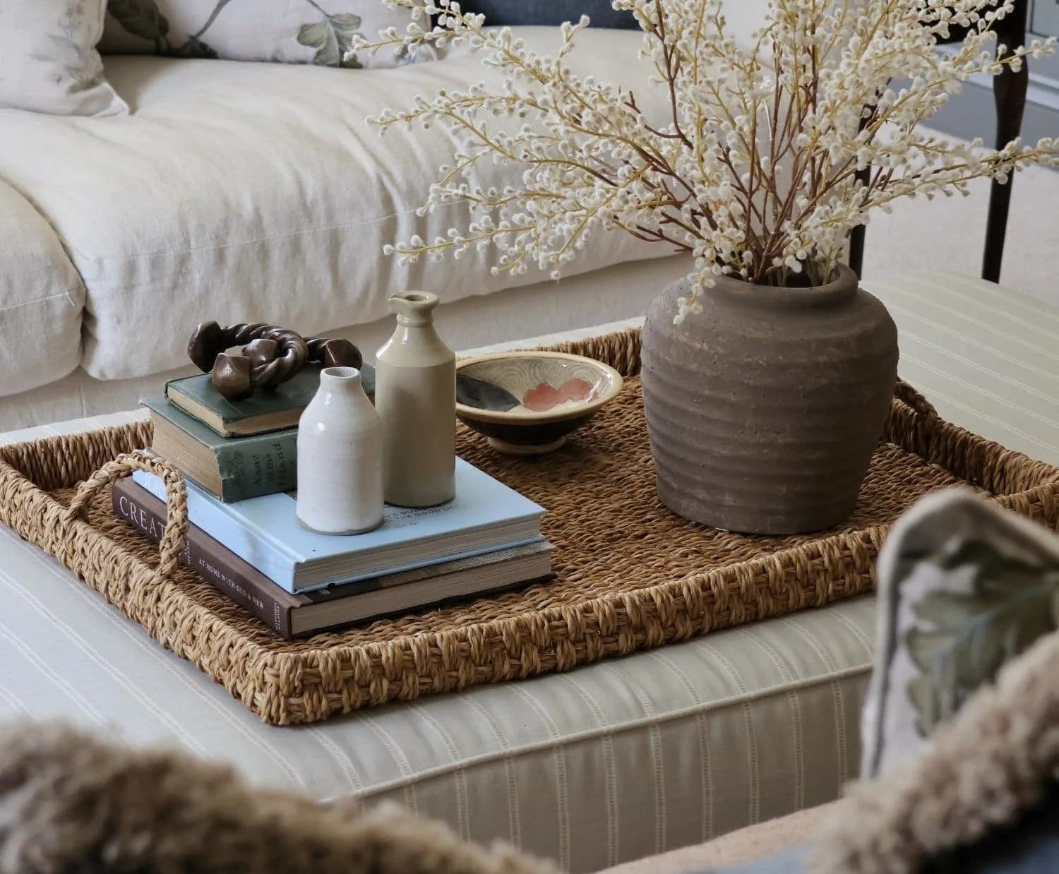 Large square seagrass tray on a striped upholstered ottoman holding a grey stoneware jar of white blossom branches, small ceramic bottles on a stack of books and a painted dish