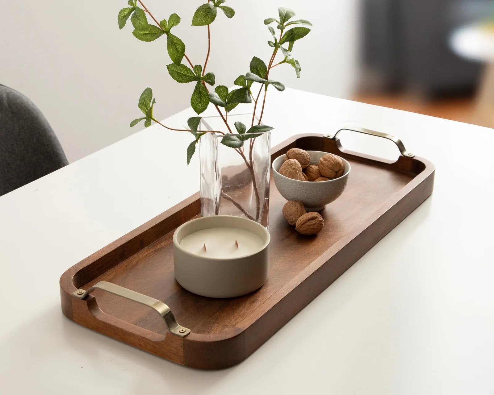 Long narrow walnut tray with brass handles on a white table, holding a glass vase with leafy branches, a wide grey candle and a small bowl of walnuts