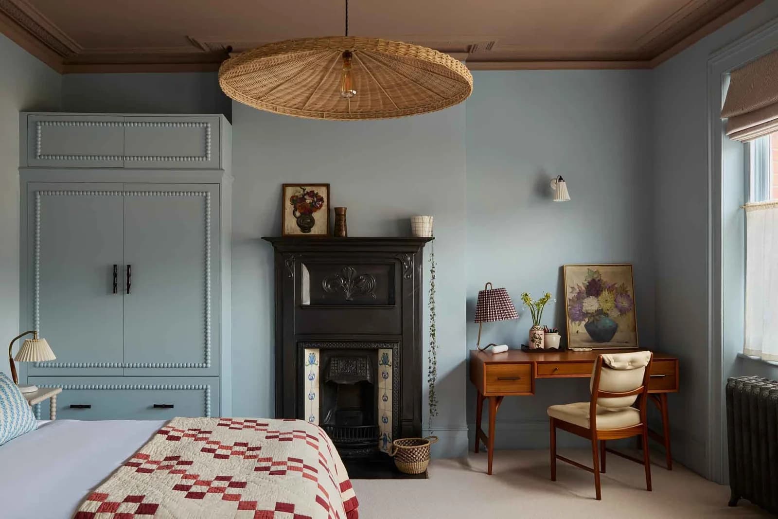 Bedroom with pale blue walls, a built-in wardrobe, window frame and radiator all painted the same blue, a mauve ceiling, a black cast-iron fireplace, a woven pendant shade and a red patchwork quilt