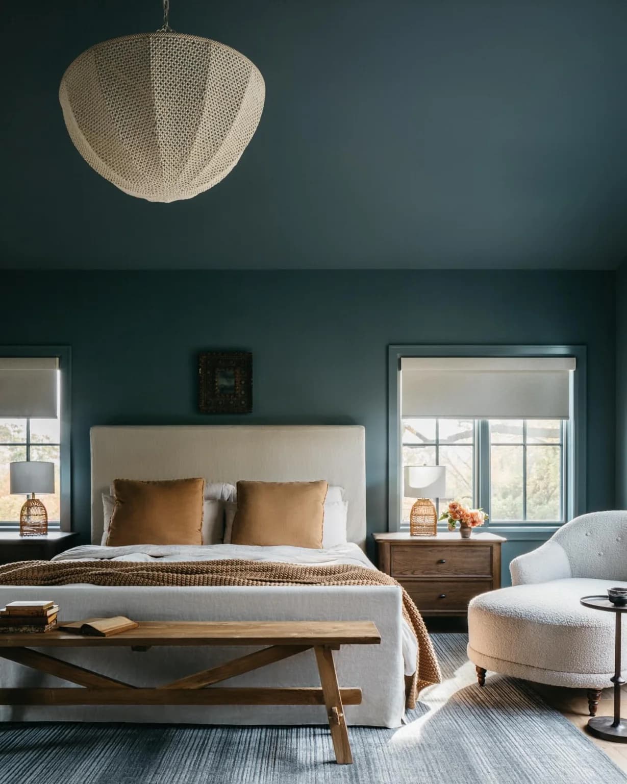 Bedroom with deep teal walls and ceiling, a cream upholstered bed with ochre pillows, a large woven pendant shade, wooden bedside tables and a wooden bench