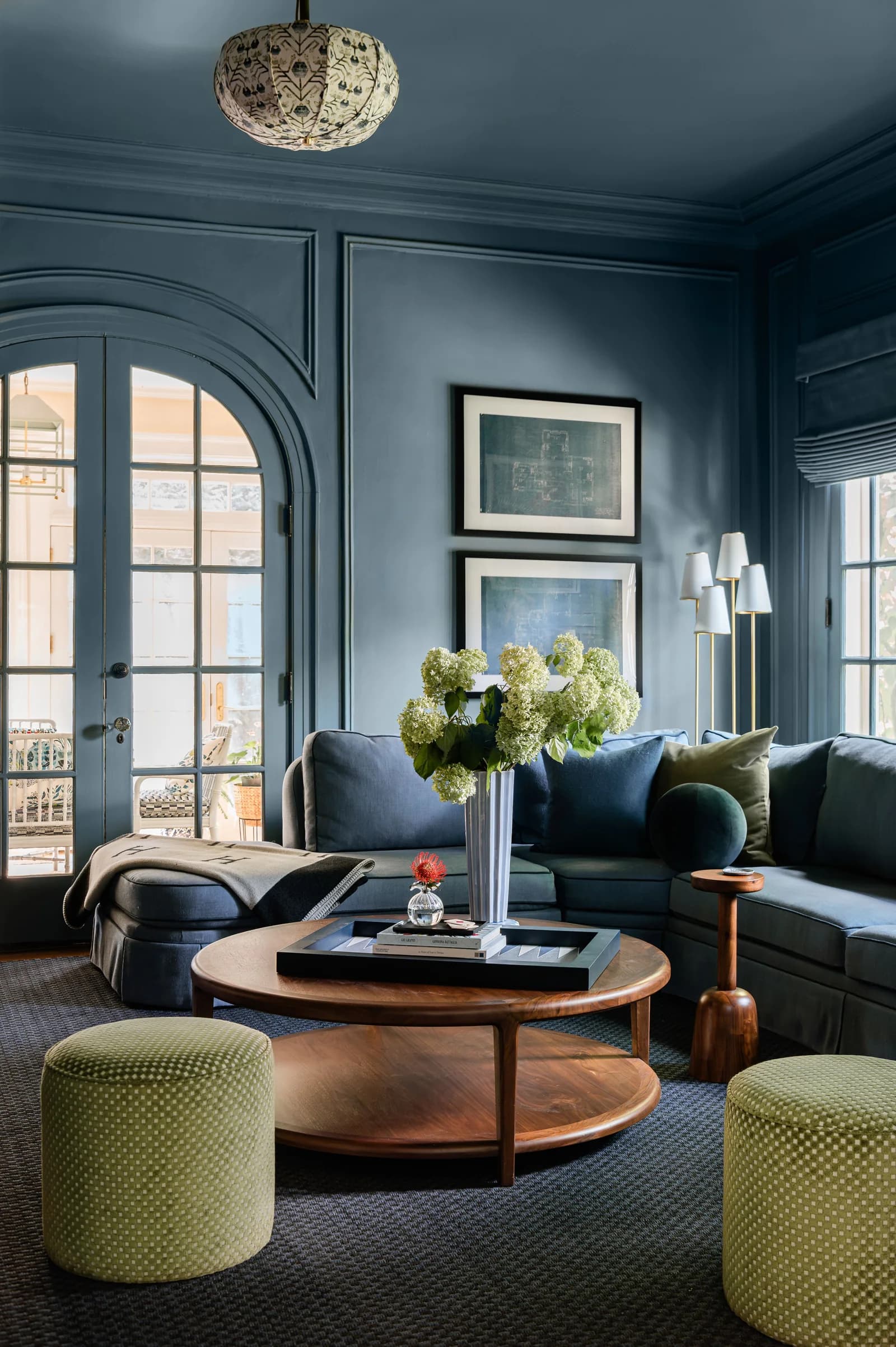 Living room with slate blue walls, ceiling, panelling and arched door frames all in one color, a navy sectional, a round wood coffee table and two green ottomans