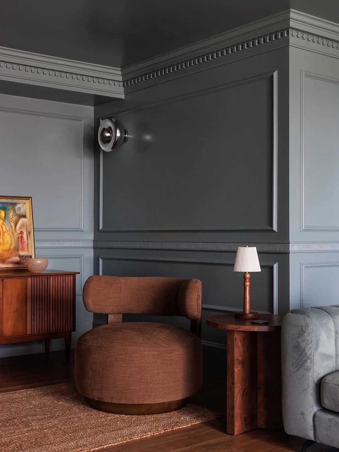 Corner of a room with grey panelled walls, cornice and ceiling painted the same grey, a rust upholstered chair, a wooden sideboard and a small table lamp