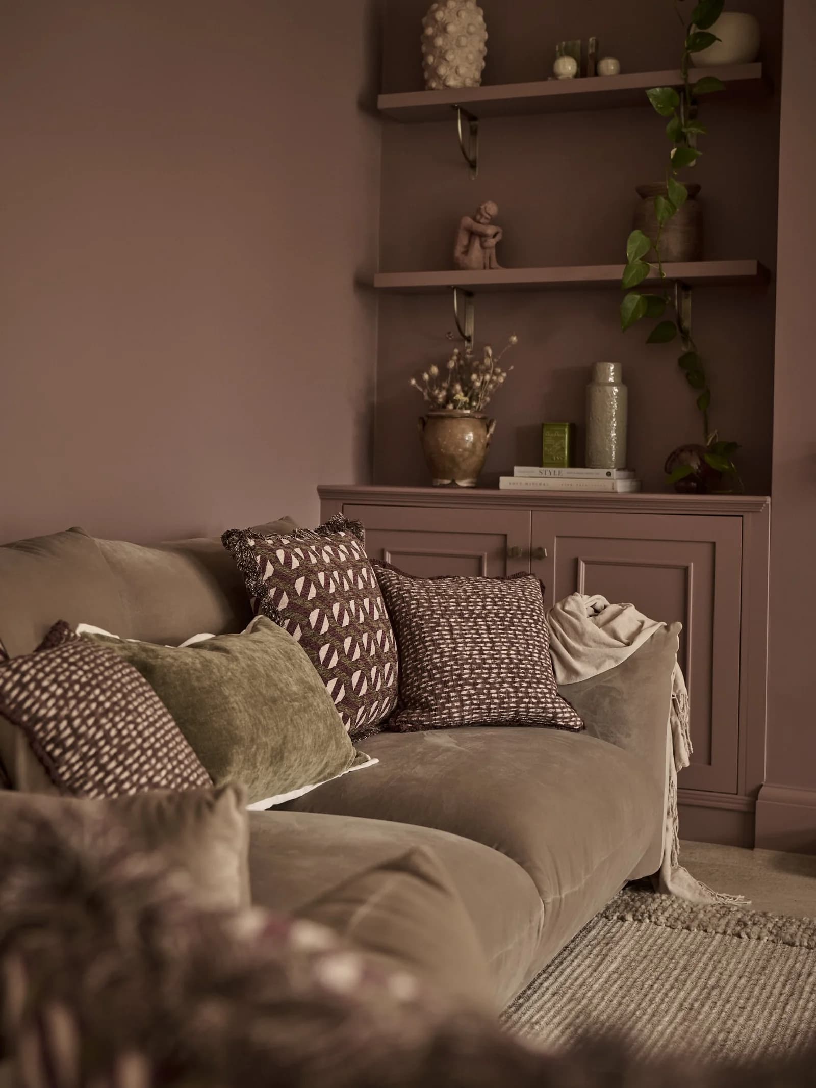 Corner of a living room with dusty mauve walls and alcove shelves and cupboards painted the same color behind a taupe velvet sofa with patterned cushions