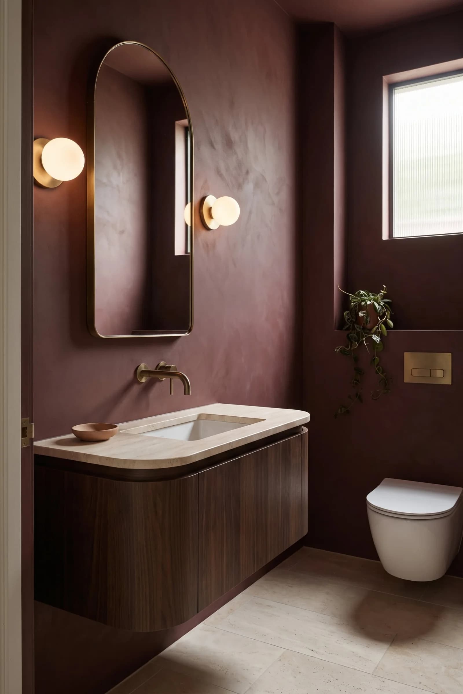 Small bathroom in a deep plum limewash-look finish on every surface, with a floating dark wood vanity, a brass wall faucet, globe sconces and an arched mirror