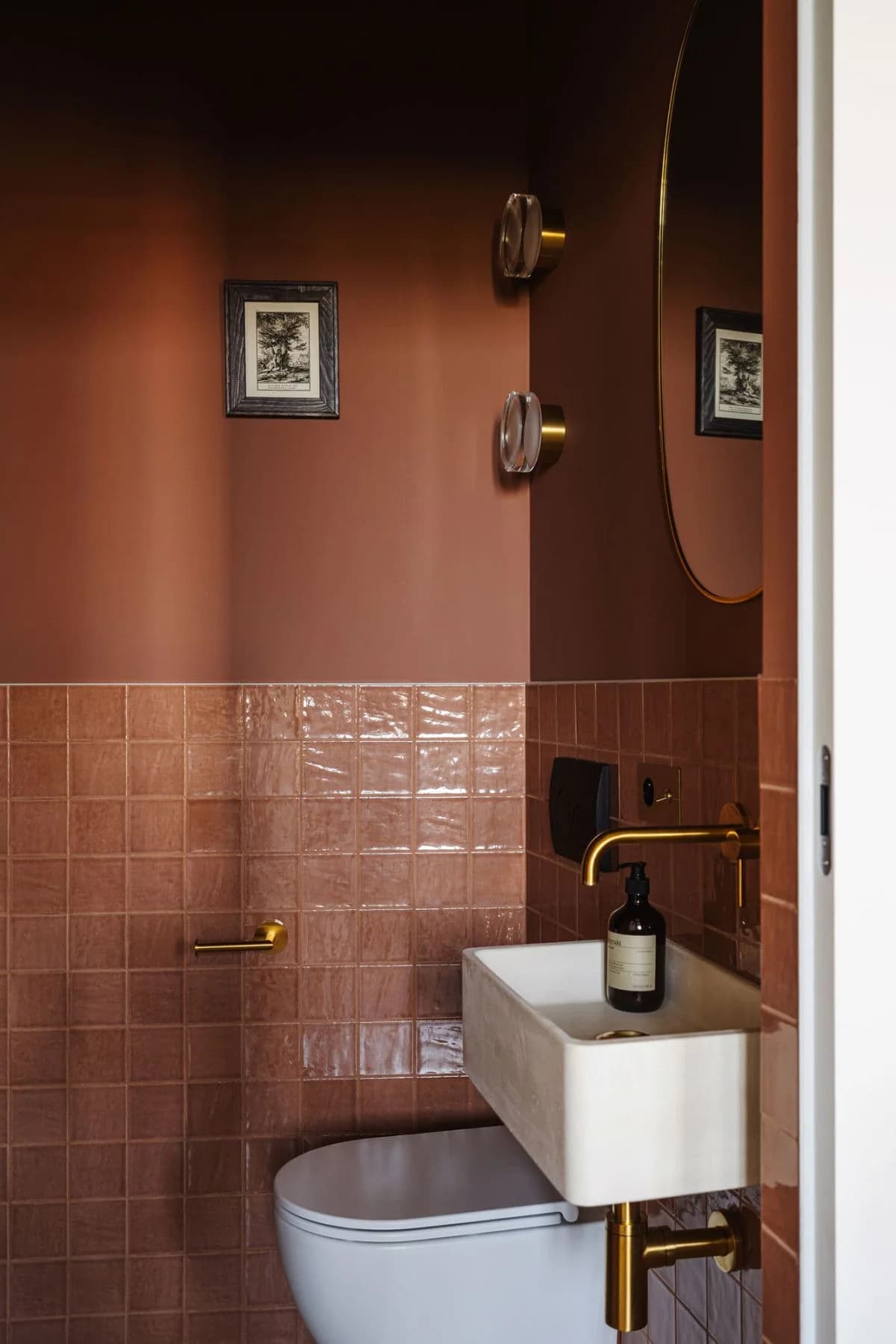 Small bathroom painted terracotta on walls and ceiling with matching terracotta square tiles below, a white block sink, brass taps and a small framed print