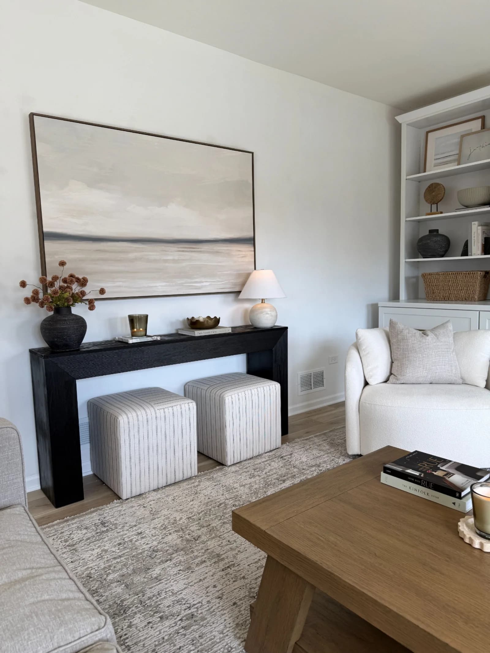 Black console with two striped upholstered cube stools tucked underneath, a small lamp, a vase of dried flowers and a wide landscape painting above