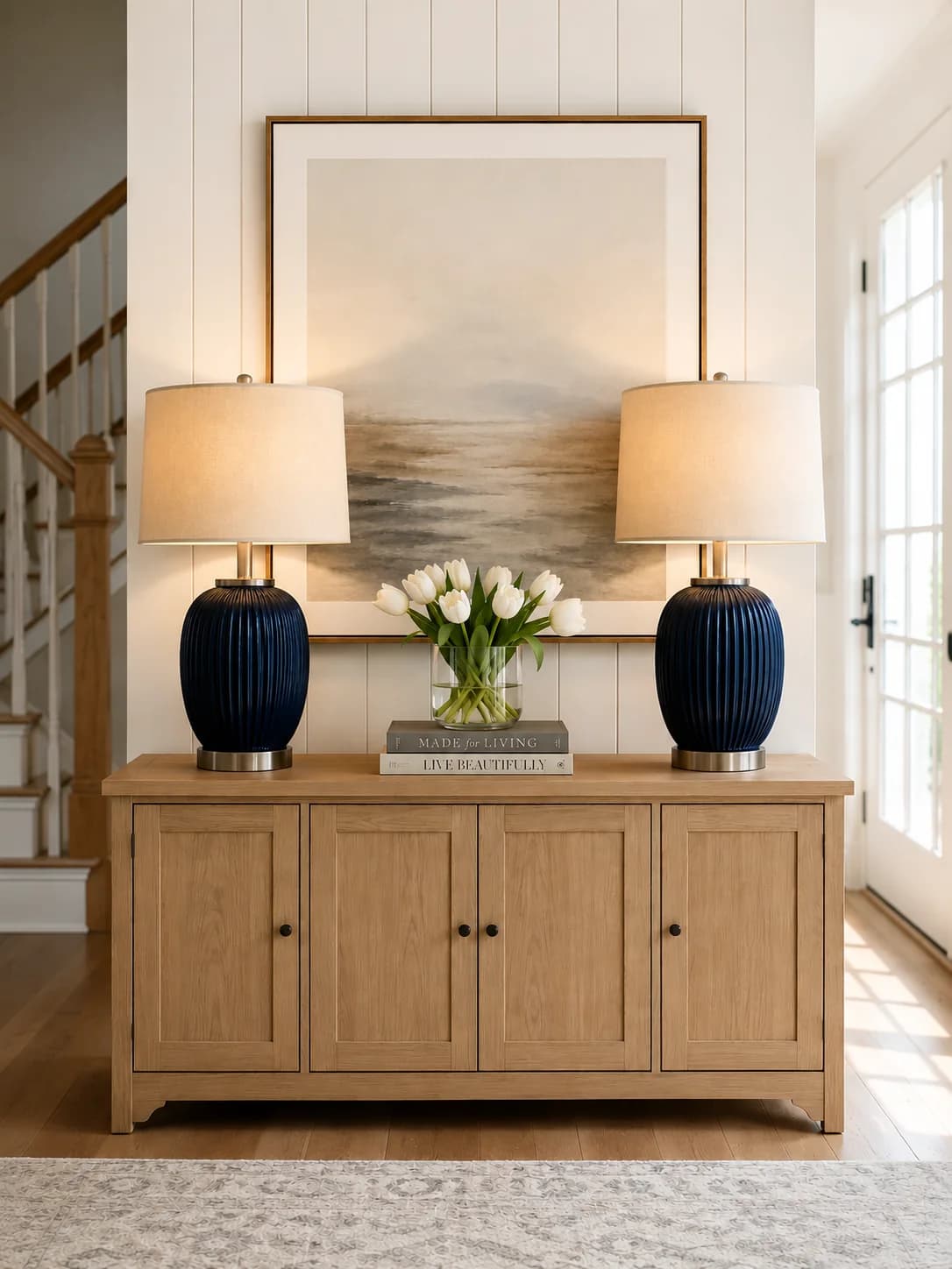 Oak sideboard with two matching navy ribbed lamps with linen shades, a vase of white tulips on a stack of books and a large seascape painting centered above