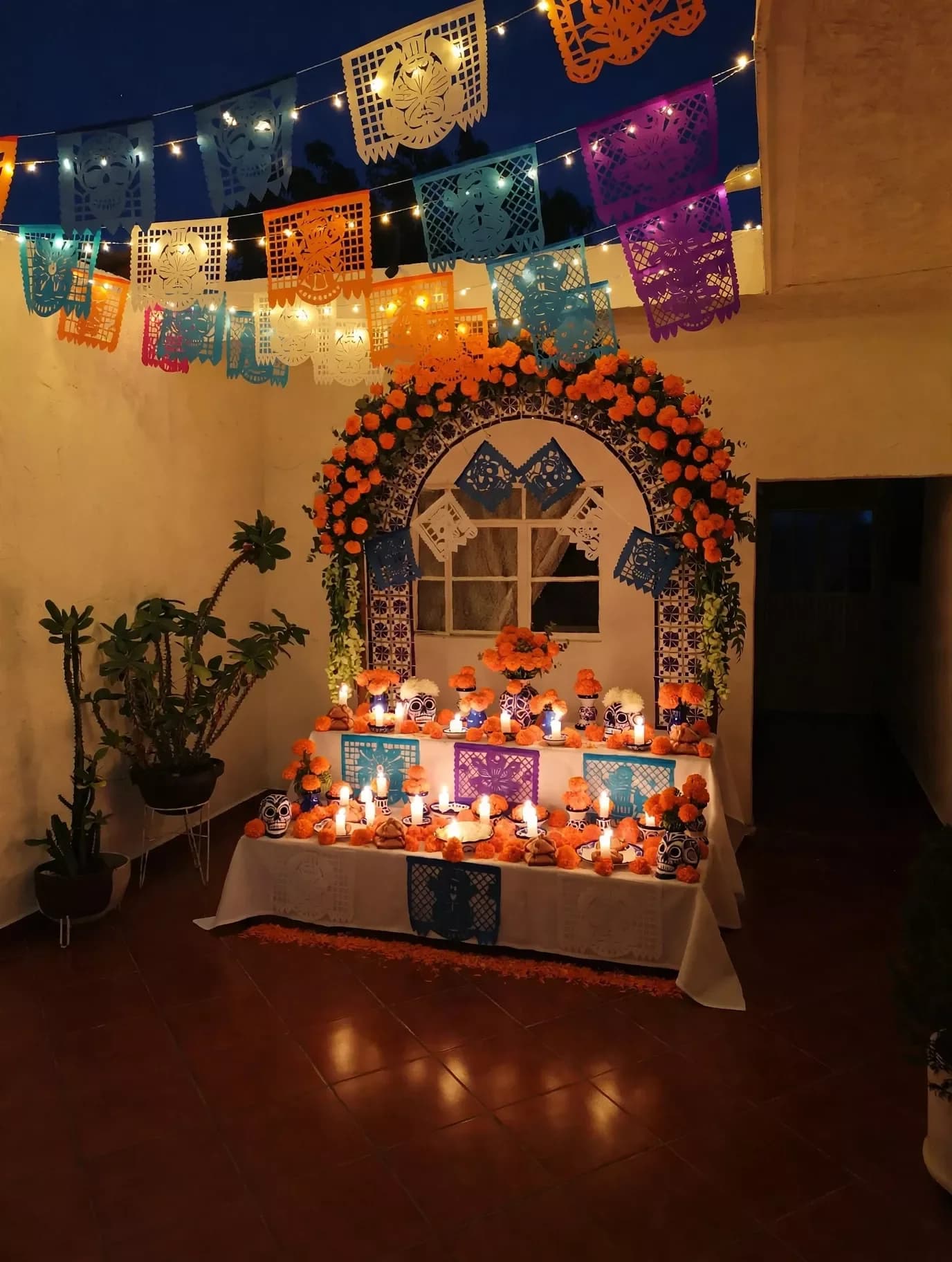 Altar de muertos con arco de flores de cempasúchil, veladoras encendidas y papel picado con luces colgado del techo en una sala a media luz