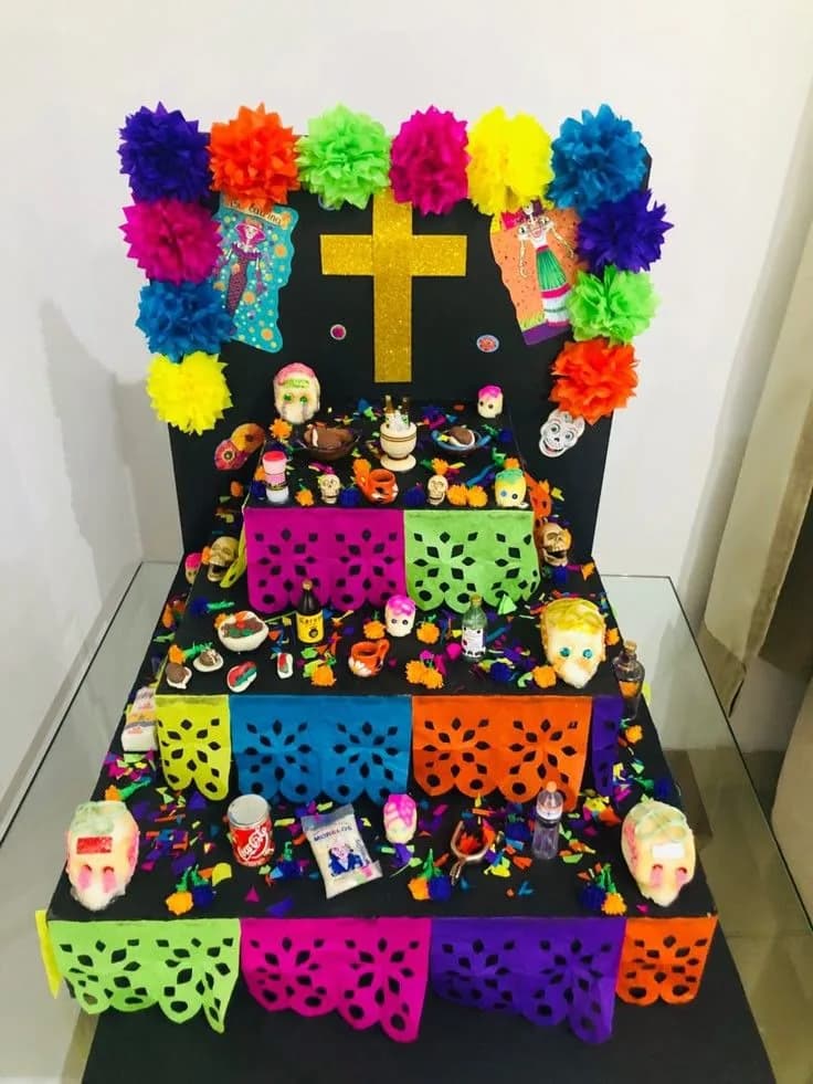 Altar pequeño de tres niveles sobre una mesa, con tela negra, papel picado de colores, cruz dorada, flores de papel y calaveritas