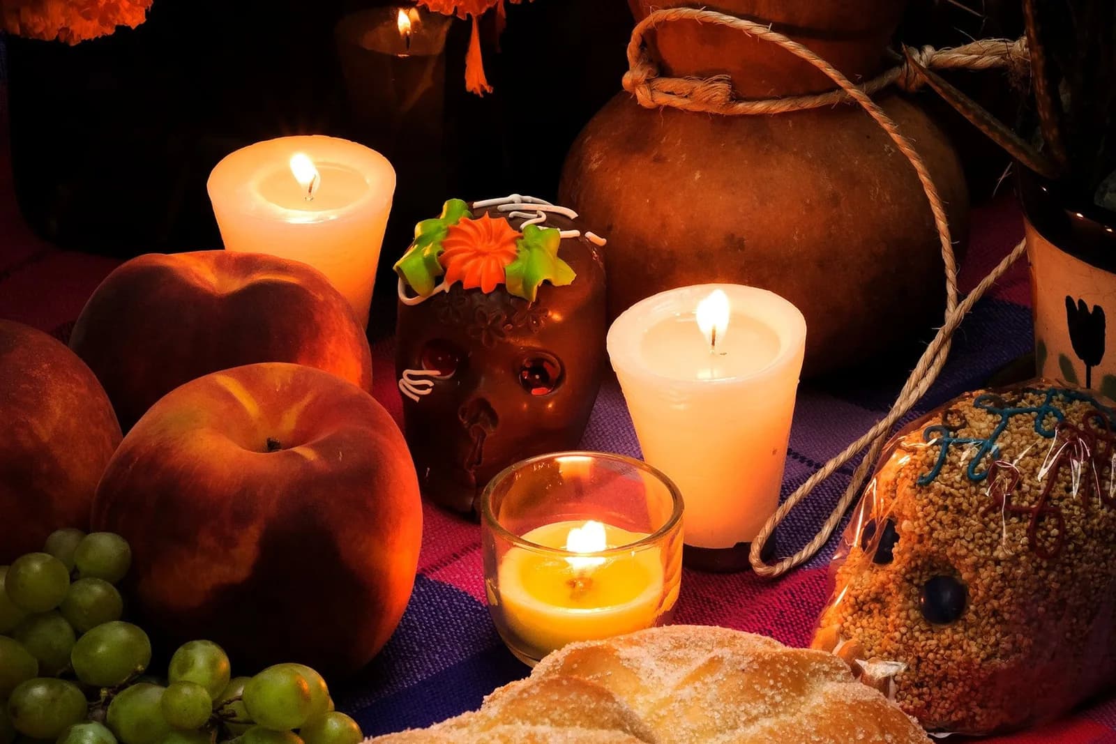 Ofrenda con veladoras encendidas, calaverita de chocolate, duraznos, uvas y pan de muerto sobre un mantel de colores