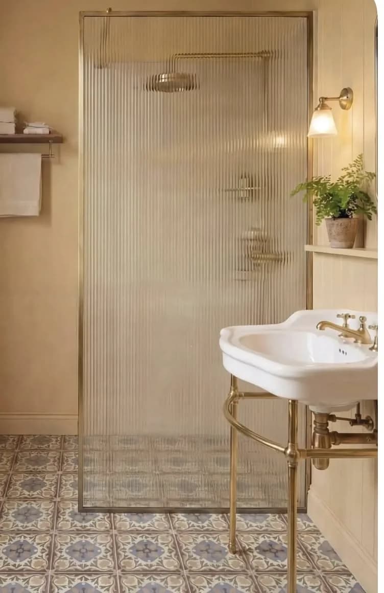 Baño pequeño color crema con ducha de vidrio acanalado, regadera dorada, lavabo de pedestal con patas doradas, aplique de pared y piso de mosaico estampado