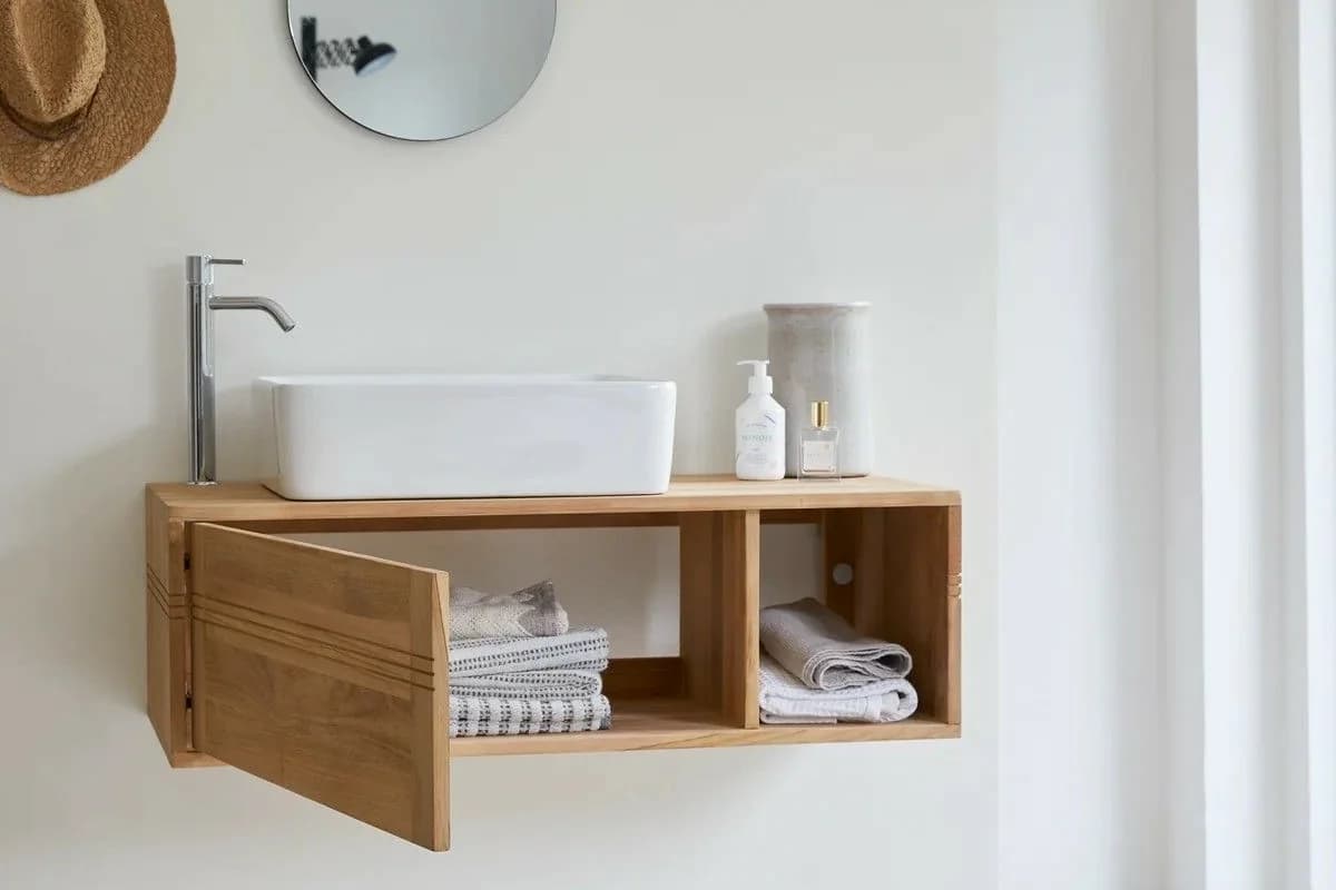 Pared blanca de baño con un mueble flotante de madera con una puerta abierta y toallas dobladas dentro, un lavabo blanco de sobreponer y un espejo redondo