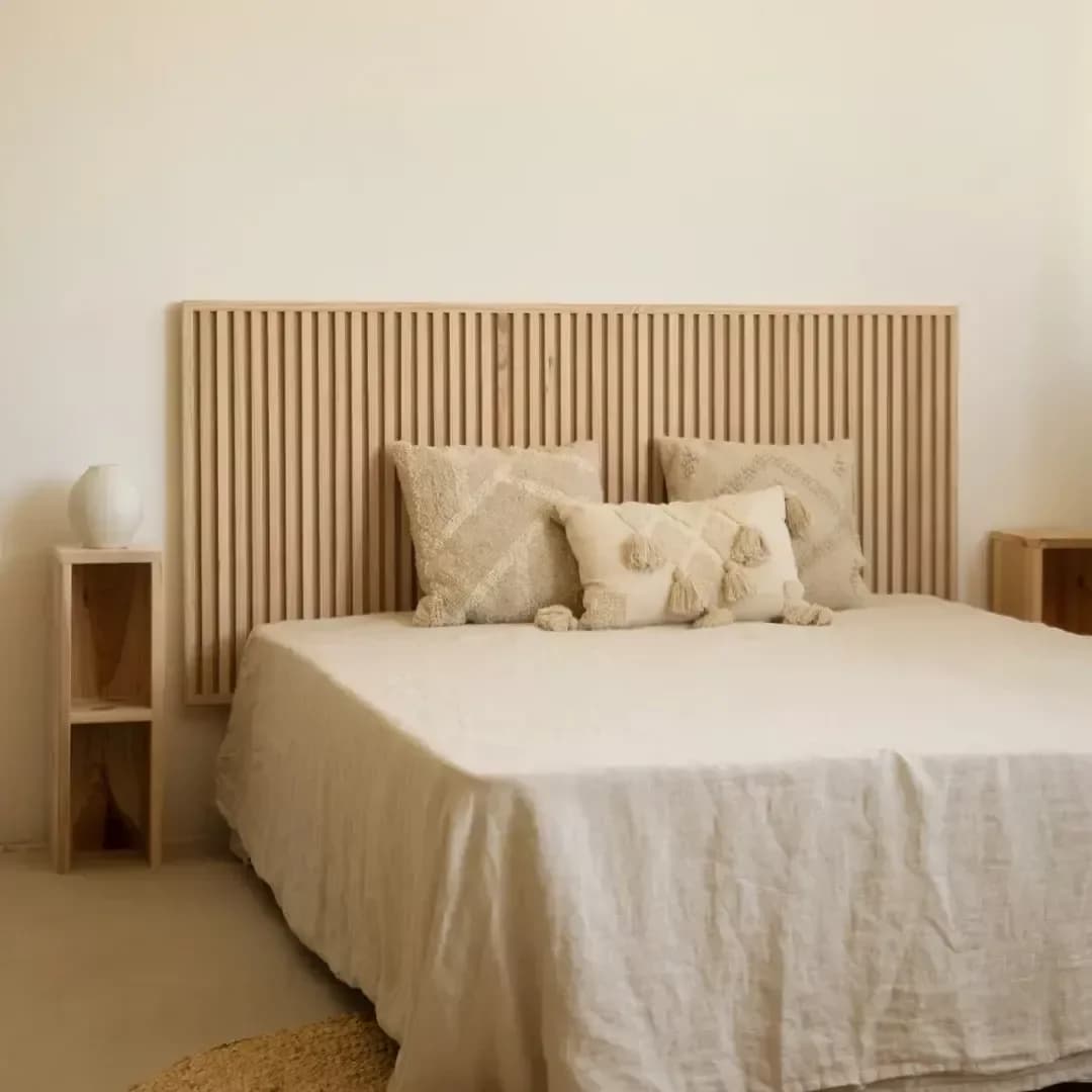 Cabecera rectangular de listones verticales de madera clara, cama con colcha de lino crema y cojines tejidos con borlas