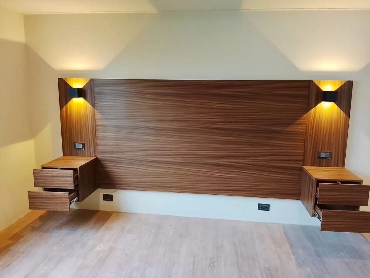 Cabecera de madera oscura fija a la pared con dos burós flotantes de cajones, contactos integrados y dos apliques que iluminan hacia arriba y hacia abajo