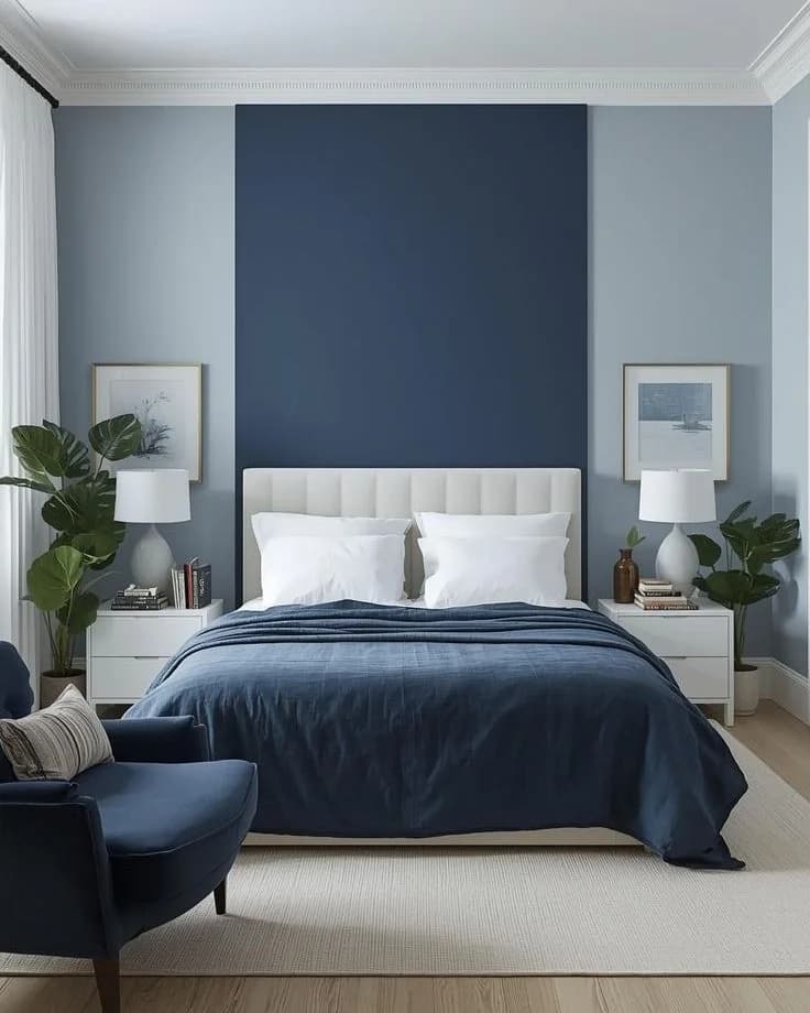 Recámara con paredes azul muy claro y una franja vertical azul marino detrás de la cama, cabecera y burós blancos, colcha azul marino y un sillón azul