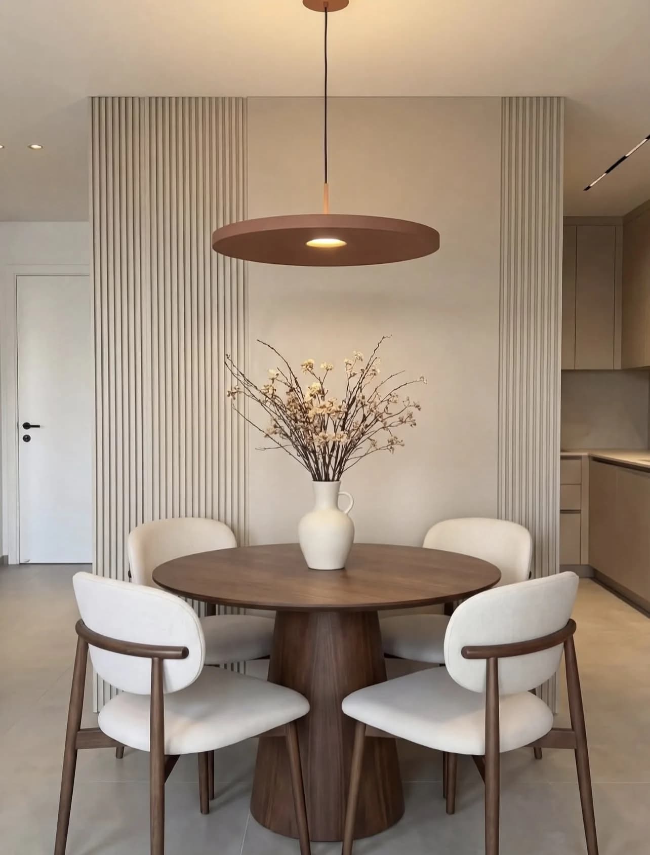 Comedor con mesa redonda de nogal de base cilíndrica, cuatro sillas blancas con brazos de madera, lámpara plana terracota y muro de lamas claras