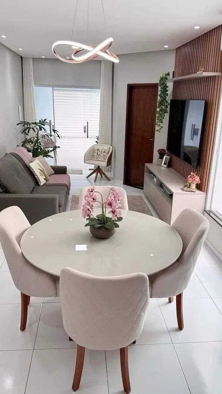 Sala pequeña con comedor de mesa redonda de vidrio, cuatro sillas tapizadas claras y arreglo de orquídeas, con sofá gris al fondo