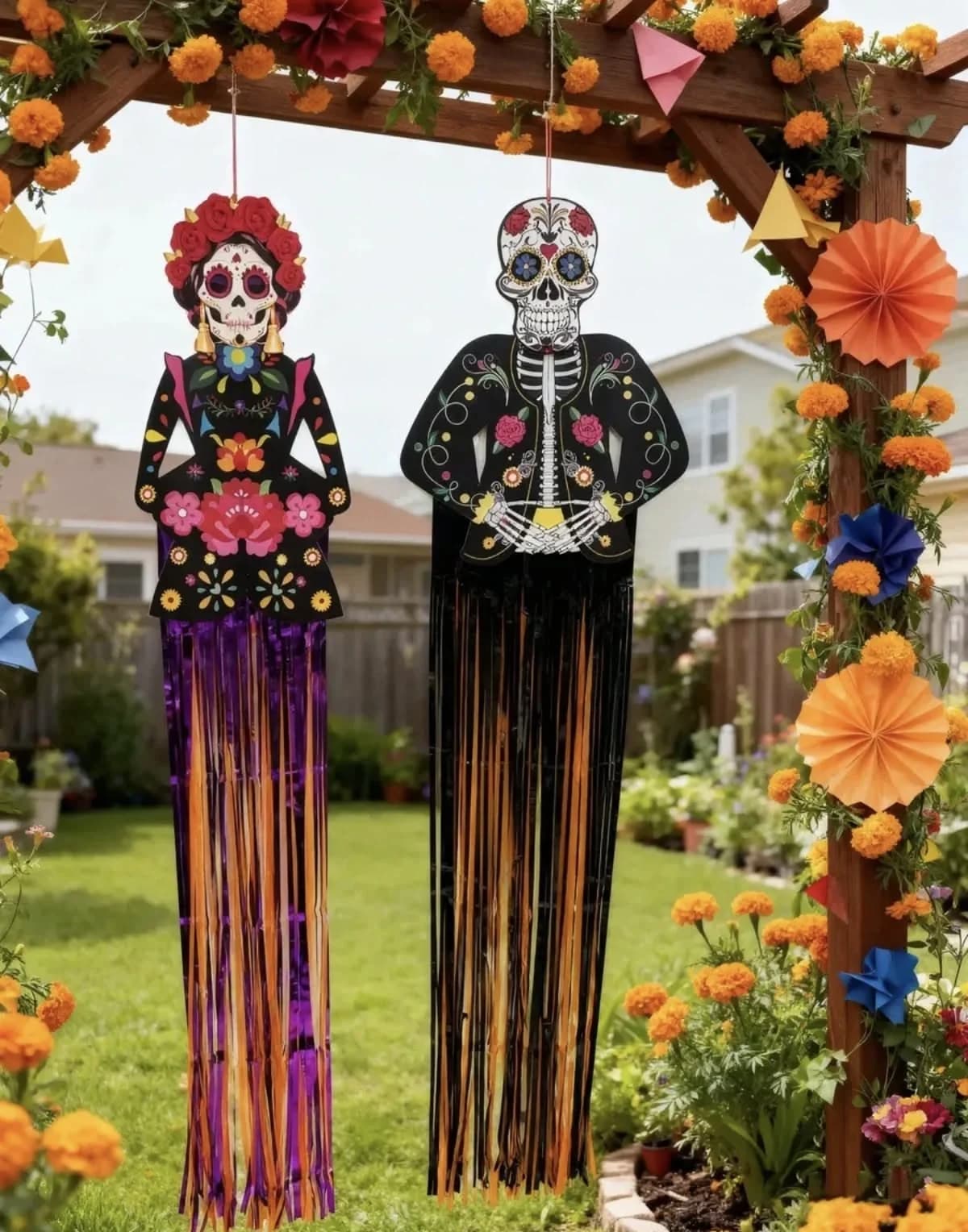 Catrina y catrín de papel con flecos largos colgados de una pérgola con flores y abanicos de papel en un jardín