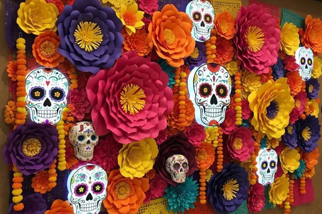 Pared cubierta de flores grandes de papel en rosa, naranja, amarillo y morado con calaveras decoradas y papel picado