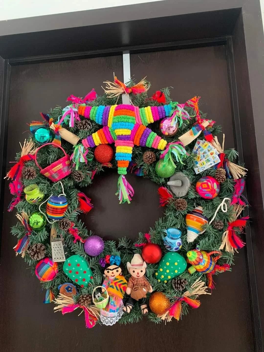 Corona navideña de pino colgada en una puerta de madera oscura, decorada con una mini piñata de estrella, esferas de colores, jarritos y juguetes artesanales