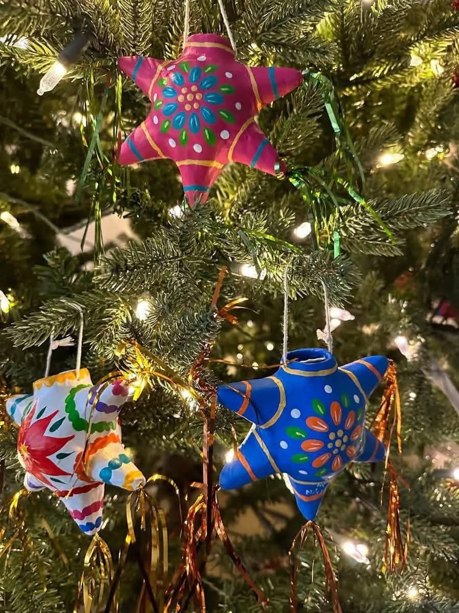 Estrellas de papel maché pintadas a mano en rosa, azul y blanco con flores de colores colgando de un árbol de Navidad con luces cálidas