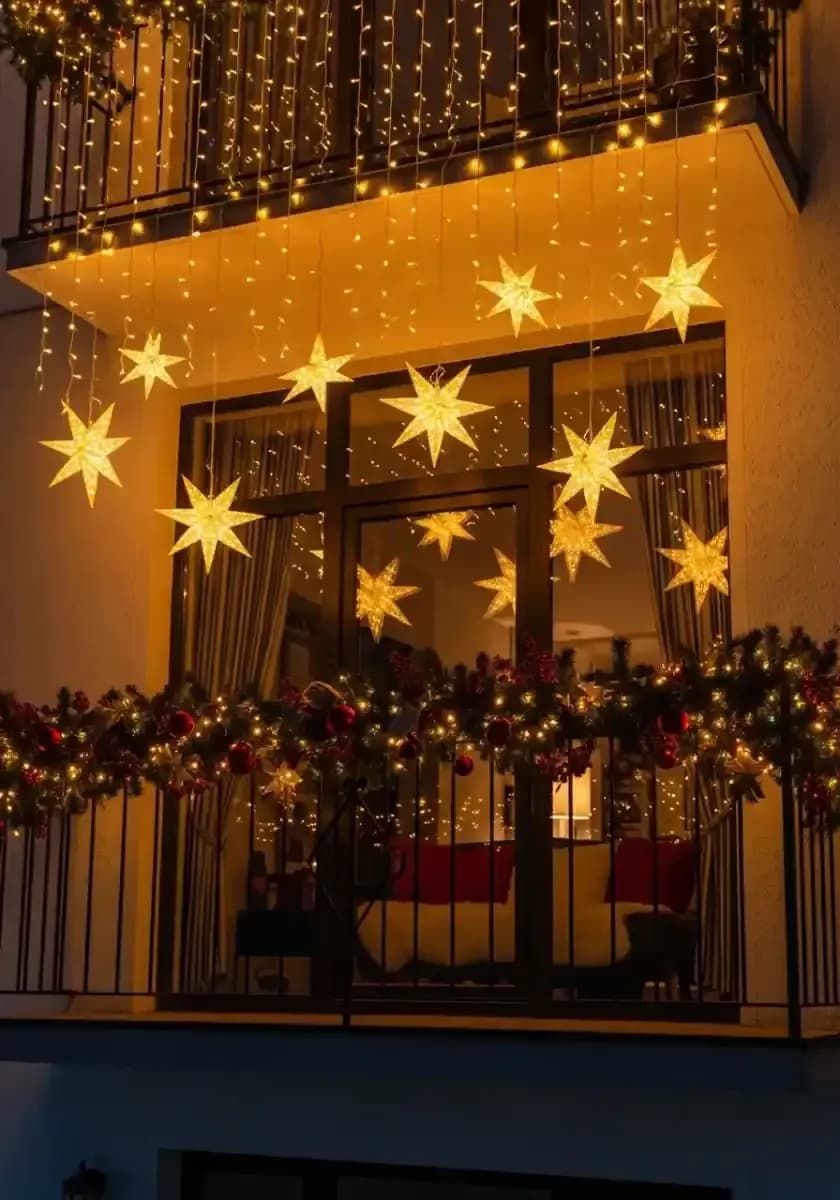 Balcón de noche con una cortina de luces cálidas y estrellas iluminadas colgando a distintas alturas y una guirnalda con esferas rojas sobre el barandal