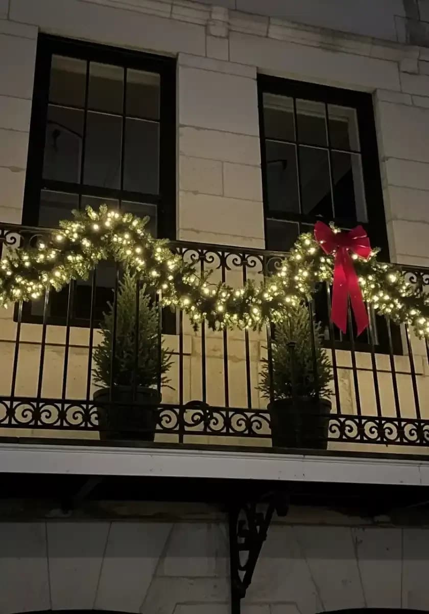 Balcón de herrería de noche con una guirnalda de luces en ondas, un moño rojo y dos pinos pequeños en maceta detrás del barandal