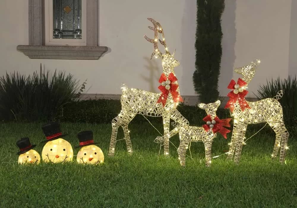 Jardín de una casa de noche con tres renos de luces con moños rojos y tres muñecos de nieve iluminados sobre el pasto