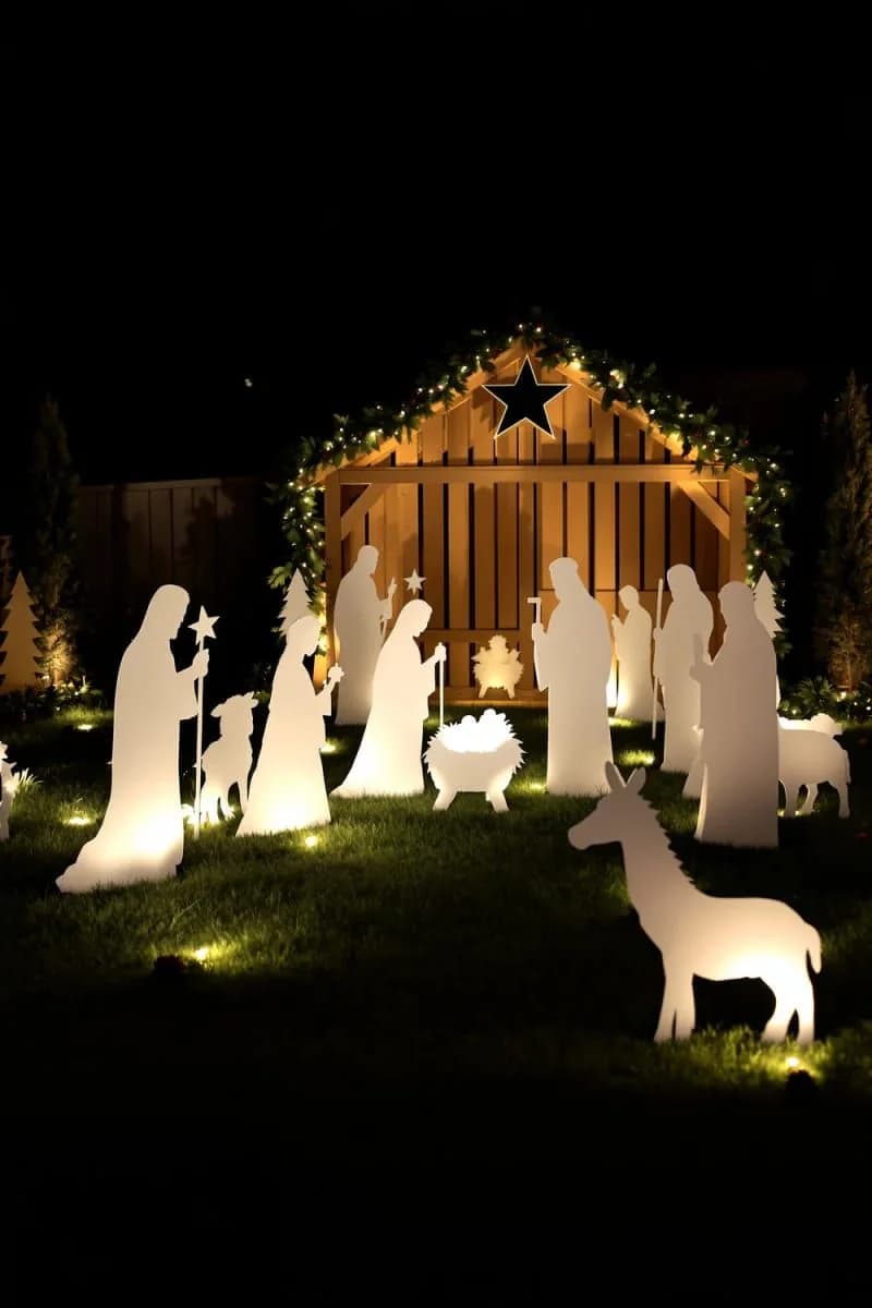 Nacimiento de exterior de noche con siluetas blancas iluminadas desde abajo frente a un portal de madera con guirnalda de luces y una estrella