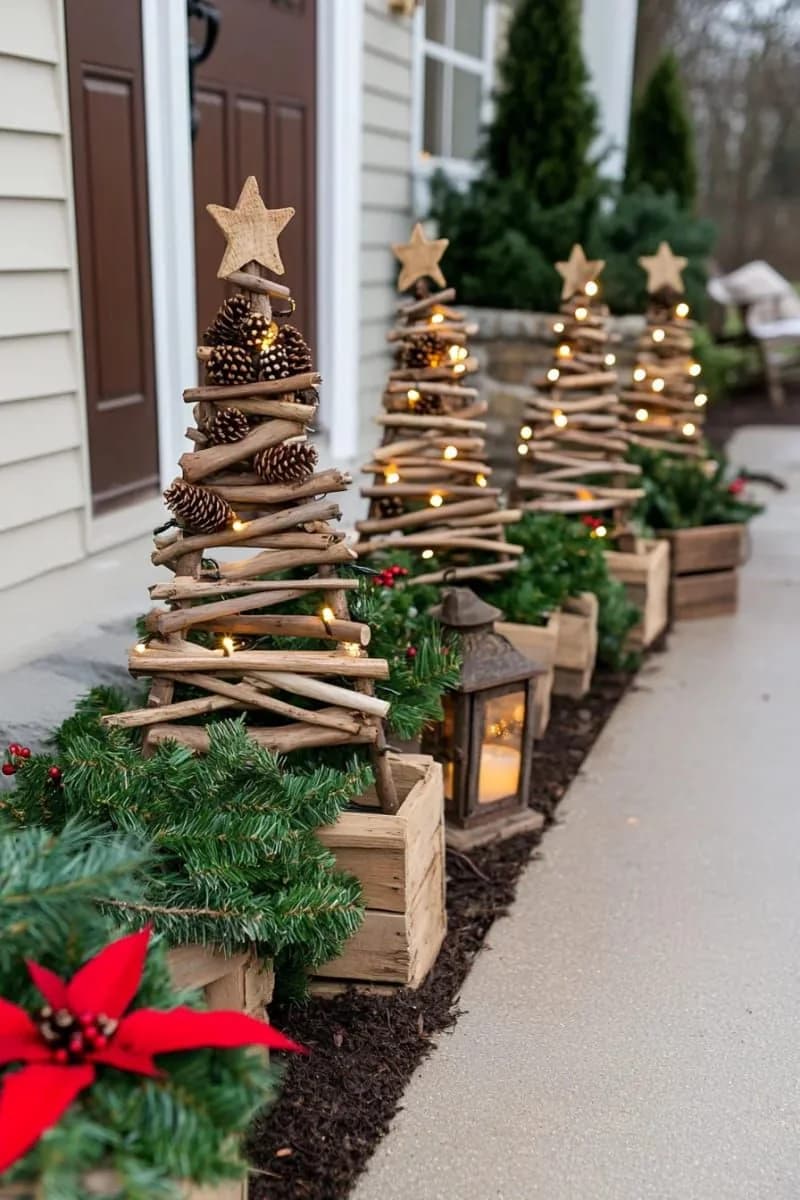 Fila de pinitos hechos con ramas secas, piñas y estrellas de madera, plantados en cajas de madera con follaje, un farol y una nochebuena junto a la entrada de una casa