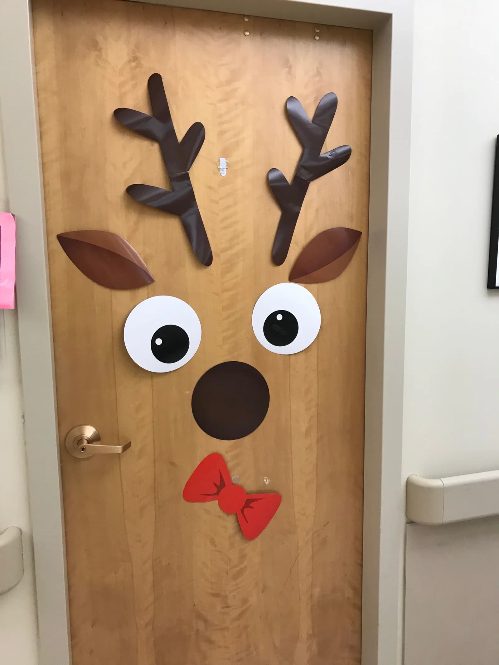 Puerta de madera clara decorada como reno con astas, orejas, ojos y nariz de cartulina y un moño rojo