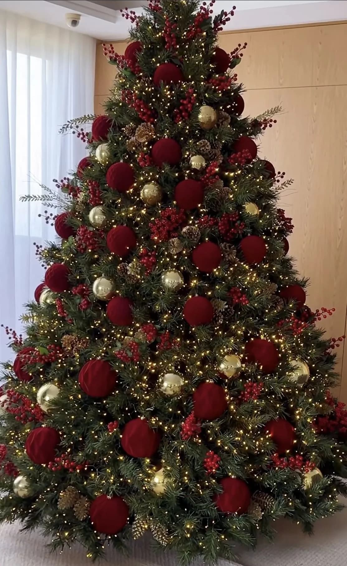 Árbol de Navidad grande con esferas mate en rojo vino y algunas doradas, muchas luces cálidas y una pared clara al fondo