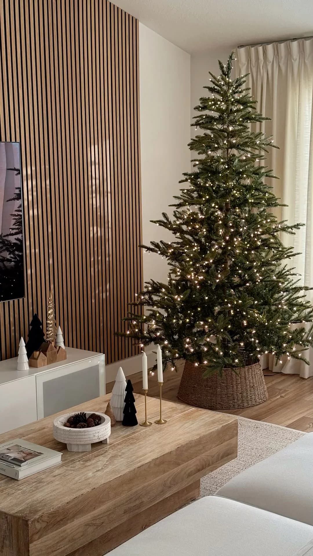 Árbol de Navidad verde sin adornos, solo con luces cálidas, dentro de una canasta de mimbre junto a una pared de lambrín de madera y una mesa de centro rústica
