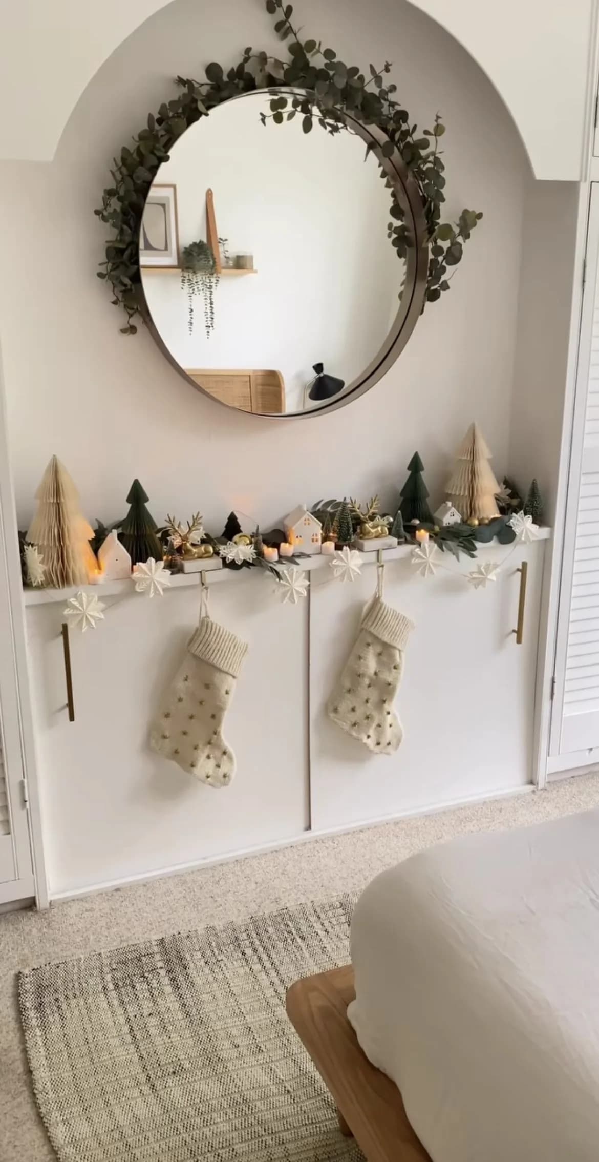 Espejo redondo rodeado de follaje verde sobre un mueble blanco con una fila de pinitos decorativos en verde y crema y dos botas navideñas de tela clara colgadas al frente